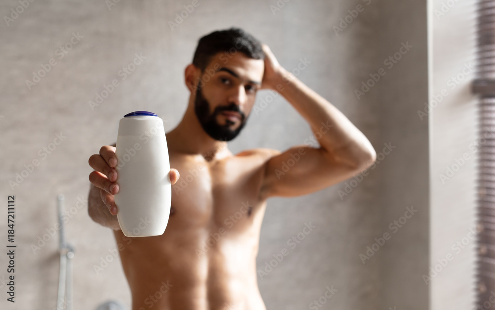 Fototapeta premium A confident Arab guy stands in a stylish bathroom, holding a shampoo bottle. He poses while raising one arm and showing off his toned physique, promoting a male cosmetic product.