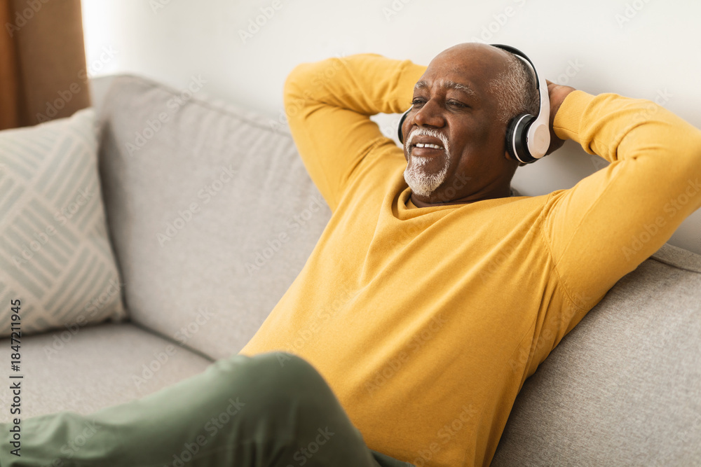 Fototapeta premium An elderly man, wearing headphones, relaxes on a comfortable sofa in his living room. He smiles as he enjoys listening to music, creating a peaceful atmosphere.