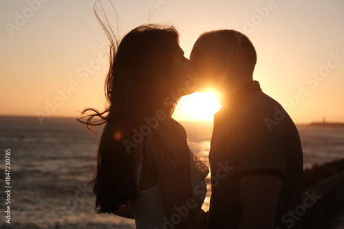 Couple sharing a romantic kiss at sunset by the sea, silhouetted against the golden light of the setting sun.