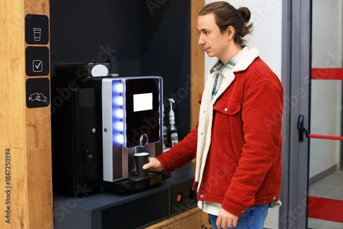  man in a red corduroy jacket using a modern, self-service coffee machine to prepare a hot beverage in an office or store.