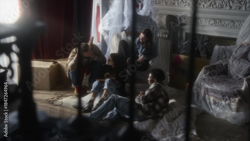 Authentic sneak shot group of young people sitting on mattresses and hanging out in secret hideout together at abandoned house with furniture in foreground, copy space