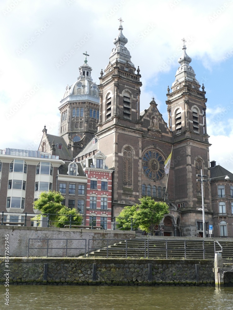 Naklejka premium Canal-side perspective revealing Saint Nicholas Basilica's ornate facade, architectural details framing historic Amsterdam urban landscape