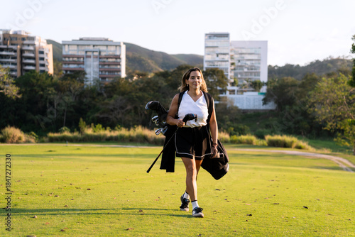 Latin hispanic woman carrying golf clubs and a bag, walking on a green golf course against a background of mountains and buildings