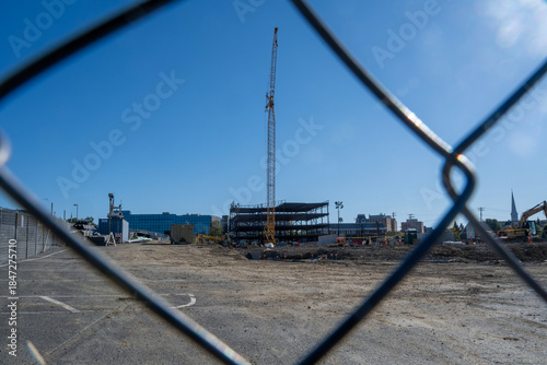 A tall yellow crane rises above a partially constructed steel-frame building, viewed through the diamond pattern of a chain-link fence. The ground is layered with dirt and gravel, scattered 