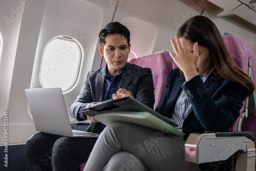Frustrated Asian business colleagues on a plane facing a stressful situation. man is explaining something with a serious expression while the woman holds her face in distress, overwhelmed by tension.