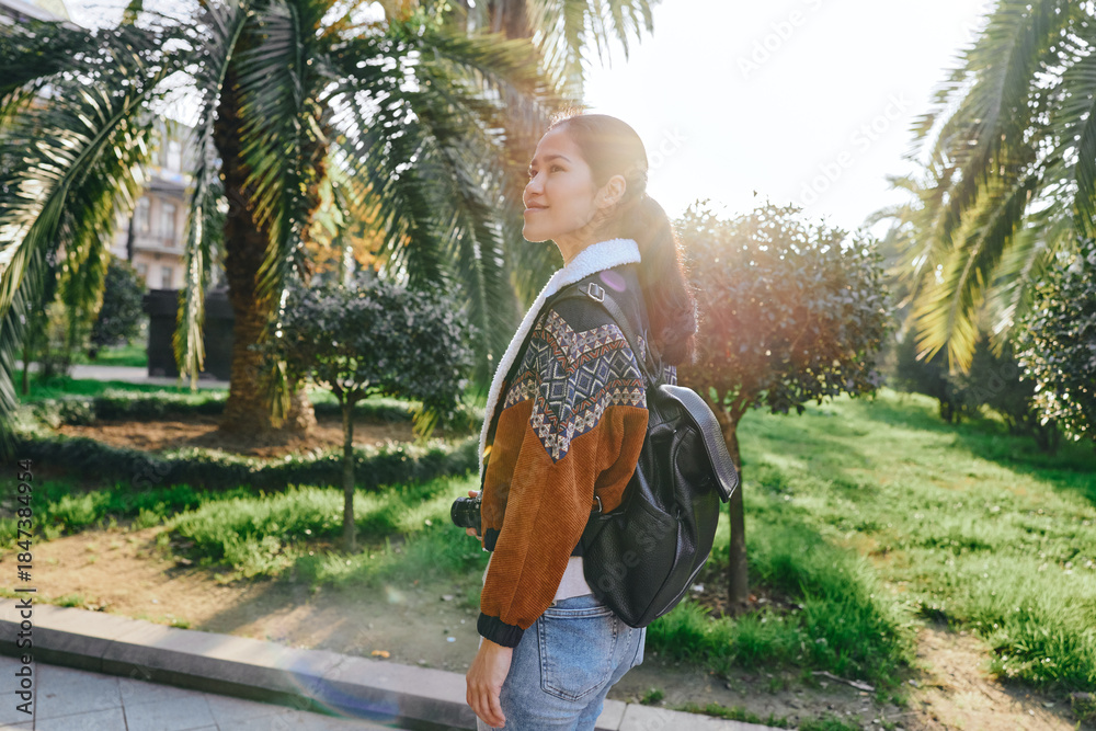 Fototapeta premium Woman with backpack walking along a sunny park path, casual outfit, palm trees in the background, natural light, candid outdoor moment, travel vibe, serene greenery