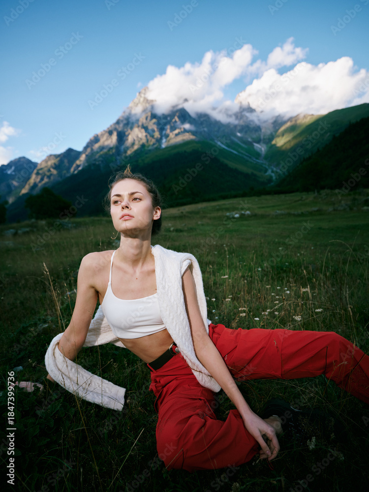 Naklejka premium Outdoor scene of a woman in a white crop top and red pants relaxing on a grassy meadow with rugged mountains and a blue sky, soft sunlight and serene alpine scenery