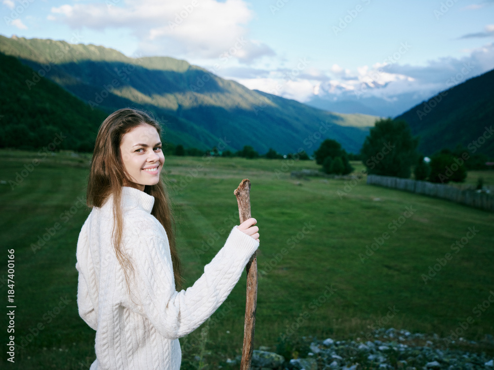 Naklejka premium Woman holding a rustic stick in a green meadow with rolling hills and distant mountains, wearing a warm sweater, bright smile, outdoor scenery and tranquil countryside.