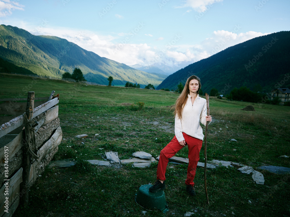 Naklejka premium Valley landscape with distant mountains, a woman in white shirt and red pants stands beside a wooden fence in a rural field, outdoors, ready for hiking and exploration