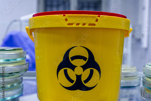A large orange container with a biohazard sign sits on a shelf among laboratory equipment and petri dishes containing bacteria.