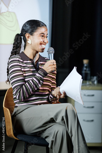 Canvas-taulu Young adult woman sitting on chair holding microphone and papers smiling while s