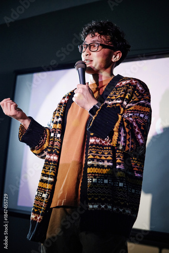 Young adult Asian man standing on stage holding microphone, speaking to audience with expressive gesture, wearing glasses, engaging in public speaking or stand up comedy performance
