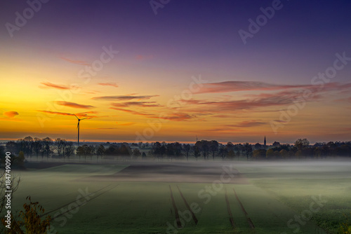 Orange, Violett und Blau fließen ineinander wie Gedanken kurz vor dem Einschlafen. Über den Feldern liegt Nebel wie ein vergessenes Versprechen, weich gezogen, fast schüchtern. Die Windräder drehen si