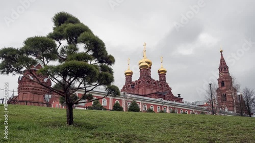 A red brick church with golden domes and a tall bell tower stands on a grassy hill under an overcast sky, surrounded by trees.