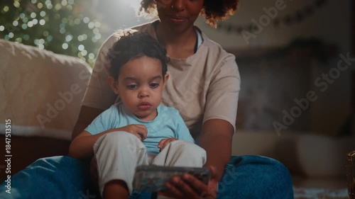 Be happy in new year and celebrate merry Christmas, african mom and son at home. Careful mother holding cute toddler and showing smartphone with cartoons, calm atmosphere of cozy home, winter holidays