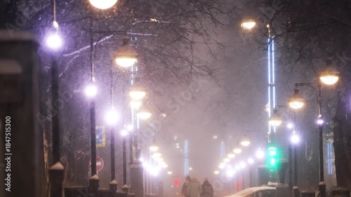 Blurry, atmospheric view of a city street during a heavy winter snowstorm at night, with streetlights glowing and cars passing by, creating a dreamy and moody urban winter scene