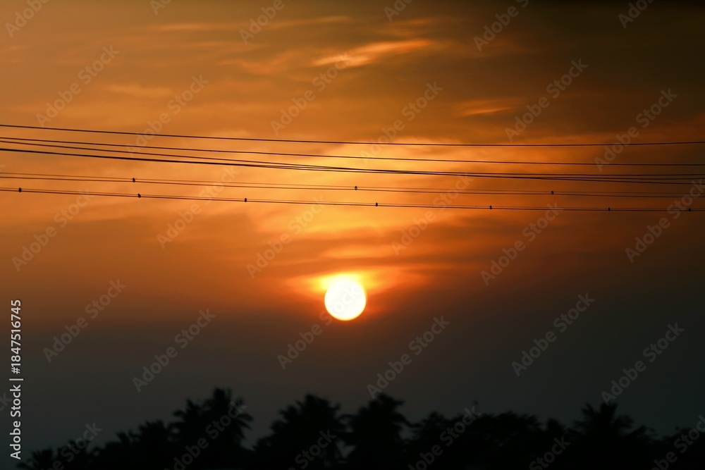 Fototapeta premium Dramatic sunset with a bright orange sun and layered clouds above a dark rural treeline, capturing calm and natural beauty