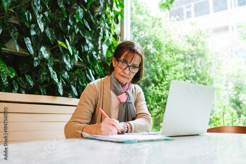 Mature woman working at a cafe with a laptop and financial documents. She reviews papers intently in a green, cozy setting, symbolizing focus, professionalism, and modern entrepreneurship.