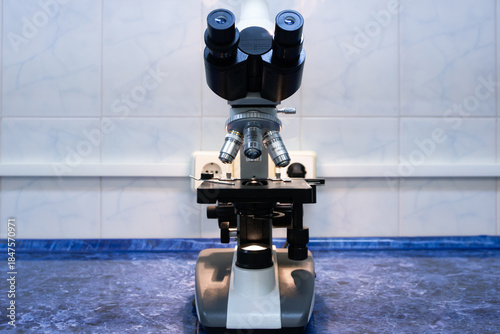 A binocular microscope with three magnification objectives stands on a workbench in the laboratory, turned on. The microscope is positioned on the table in working order.