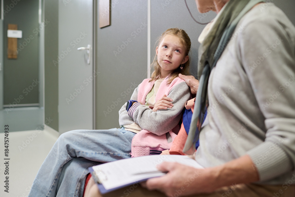 Obraz premium Caucasian girl sitting with arms crossed next to senior Caucasian woman holding clipboard, both waiting in hallway, girl looking up attentively at woman, casual setting