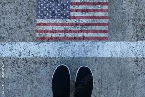 Symbolic image of a man shoes on asphalt road border, USA flag on the other side. USA immigration law and travel concept. 