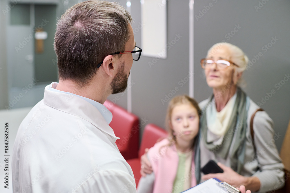 Obraz premium Caucasian male doctor speaking with senior Caucasian woman and young Caucasian girl in medical office, senior woman holding smartphone while girl standing close to her