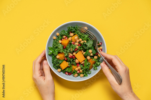 Healthy Kale Salad with Chickpeas, Pumpkin, and Pomegranate Seeds in Female Hands on Yellow Background top View