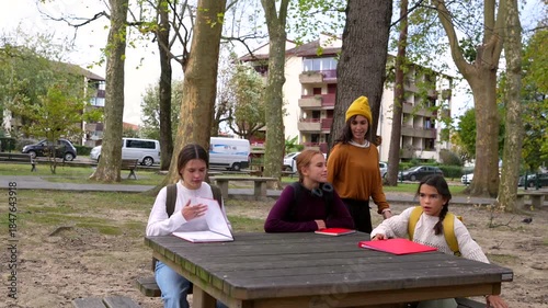 Young female students sitting down at a wooden picnic table in a city park, opening their notebooks to start studying and discussing their homework in a cheerful and collaborative manner