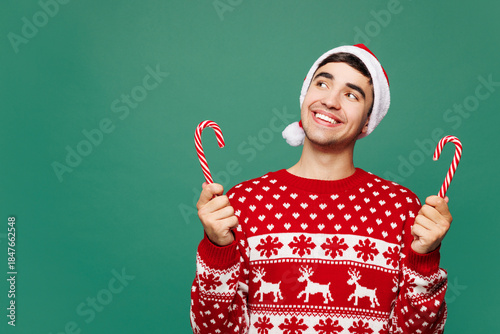 Merry young man wear red warm cozy Christmas sweater Santa Claus hat posing hold in hand candy cane look aside on area isolated on plain green background. Happy New Year celebration holiday concept.