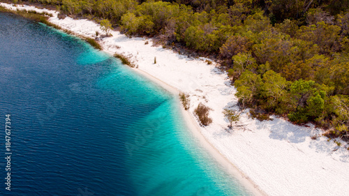 Aerial view of the crisp white sands meeting the deep azure waters of Lake McKenzie, fringed by emerald forests, creating a serene contrast, Lake McKenzie, Queensland, Australia.