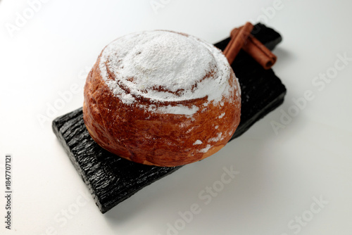 Round, delicious bun with cinnamon and powdered sugar on a black wooden board on a white background