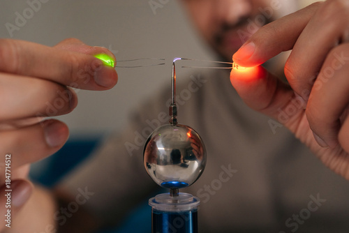 Close-up hands of scientist holding two leds near mini tesla coil, observing electrostatic discharge and electrical sparks illuminating light-emitting diodes during physics experiment, closeup.