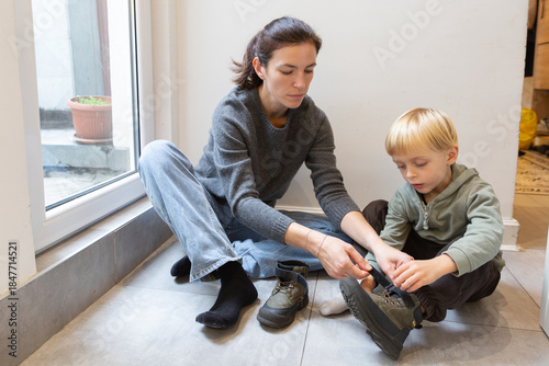Fotografía Woman assisting young boy with putting on shoes, sitting on floor near window, s