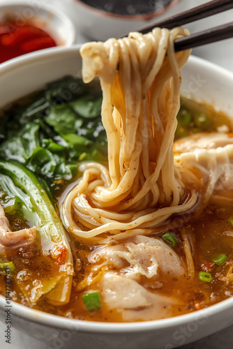 Close-up view of clear chicken broth noodle soup featuring handmade chicken wonton dumplings and fresh green bok choy. A small dish of chili oil and soy sauce is placed next to the