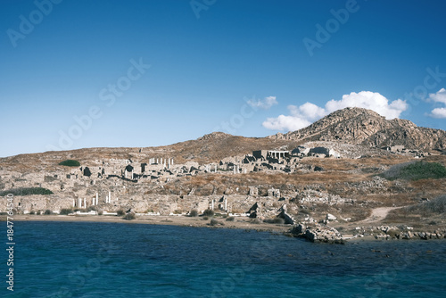 Clear coastal view of Delos island showing scattered ancient ruins on a rocky hillside, bright blue sky, and deep turquoise water below.