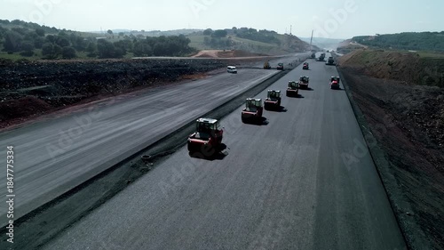 Aerial view of asphalt road construction with multiple road rollers compacting fresh pavement. Modern highway development, transportation infrastructure, civil engineering, and large-scale constructio
