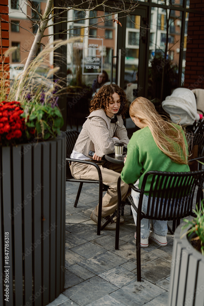 Naklejka premium Two young women enjoy a relaxed moment together at a charming cafe patio. Surrounded by vibrant flowers, they share laughter and conversation. The scene is filled with warmth and friendship.