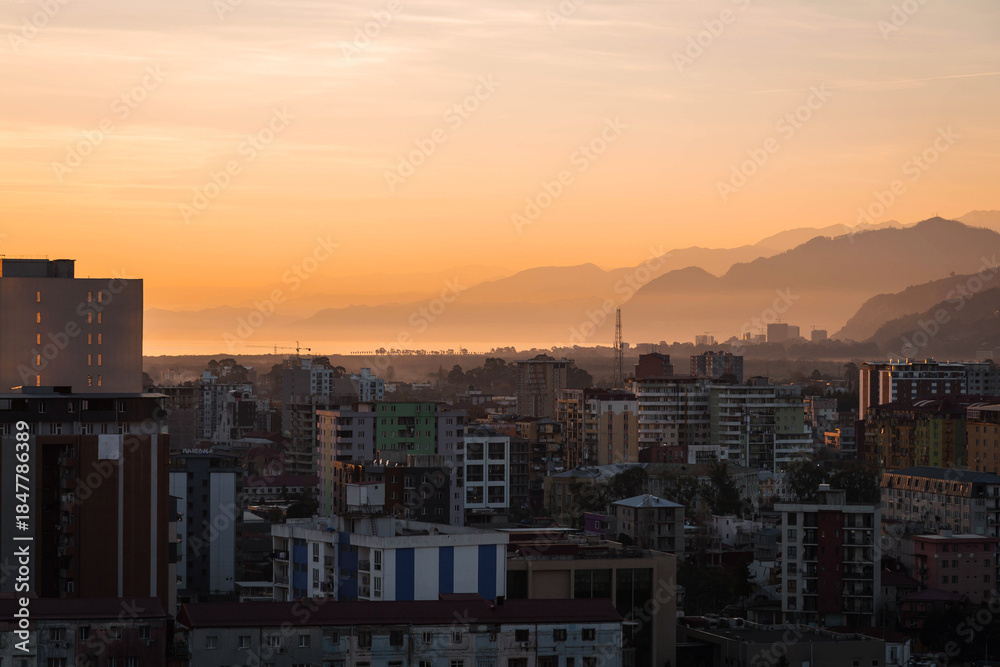 Fototapeta premium Urban cityscape at sunset with warm golden sky, modern residential buildings, layered mountains in background, evening atmosphere, calm mood, travel and real estate concept