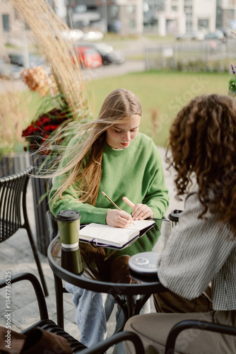 Canvas Print Two friends sit at a small table in an outdoor cafe, one writing in a notebook while the other enjoys a drink