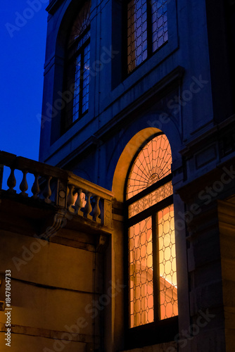 Warm light glowing through historic arched windows at blue hour, revealing elegant architectural details, stone textures and a quiet urban atmosphere shaped by contrast, depth and evening calm