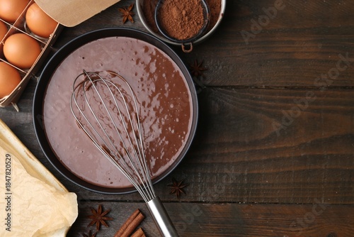 Liquid chocolate dough in bowl, whisk and ingredients on wooden table, flat lay. Space for text