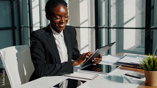 Young adult black businesswoman focusing on her work, expertly using a digital tablet while sitting at a modern office desk bathed in natural sunlight
