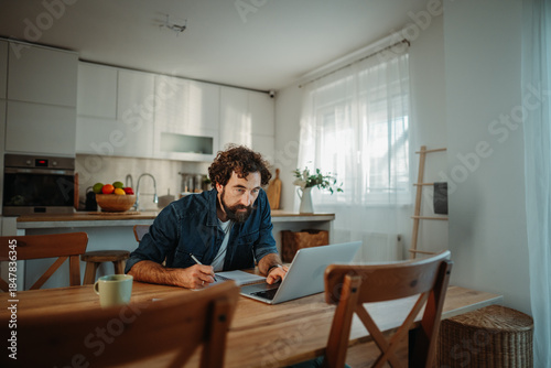 Man working remotely using laptop and notepad at home kitchen table