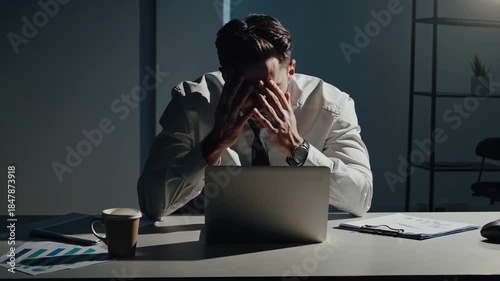 Businessman sitting at an office desk in dark lighting, holding his head in his hands, feeling stressed and overworked from a deadline while working late