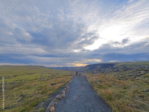 Two hikers walking along a gravel path in the vast, rugged Icelandic highland landscape at sunset. The trail is bordered by mossy green and brown fields under a dramatic, expansive sky.