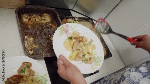 Close up of hands presenting white decorative plate with fried meat and potato slices, with prepared dishes visible on counter in background. Pour fried potato to plate.