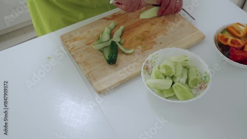 Close up of hands wearing a yellow green apron cutting fresh green cucumber into strips on wooden board with knife, put to bowl, preparing healthy vegetables for cooking meal at home.