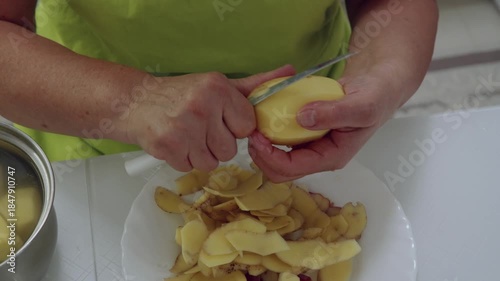 Close up of hands in yellow green apron peeling raw potato, wash in pan and put onto white decorative plate, organizing prepared vegetables for cooking meal at home.