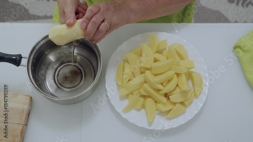 Close up of hands in yellow green apron cut and transferring peeled raw potato slices from stainless steel bowl to white decorative plate, arranging ingredients for cooking meal at home.