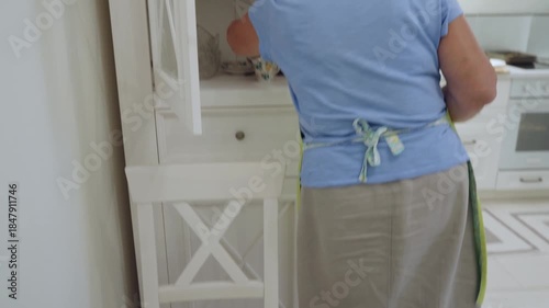 Close up of hand in yellow green apron arranging white floral patterned teacup with saucer on yellow green placemat, preparing elegant table setting in modern kitchen interior.